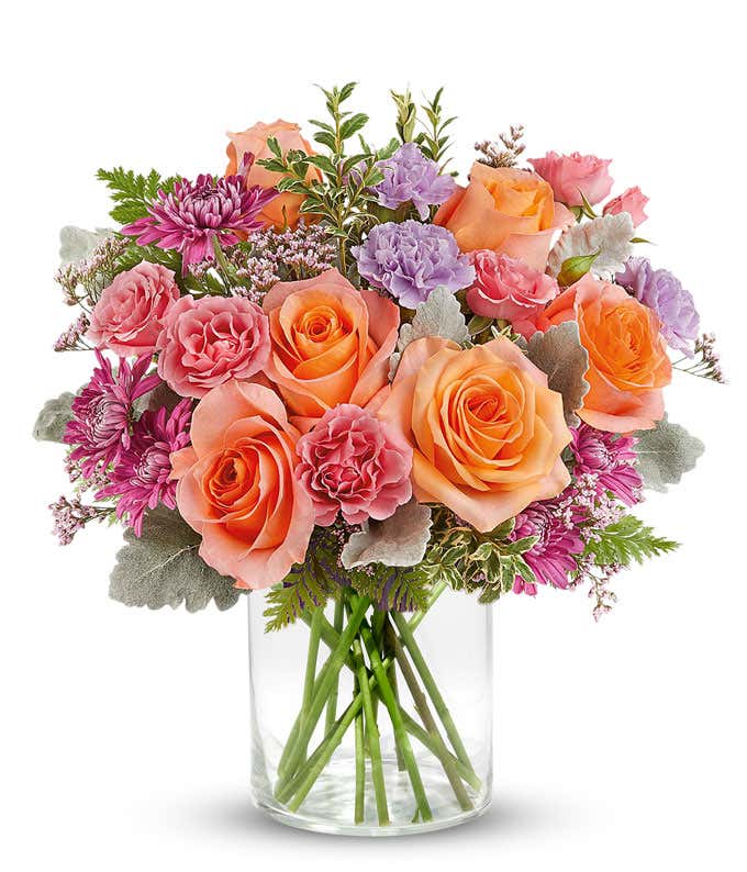 Vibrant floral arrangement featuring orange roses, pink miniature carnations, lavender carnations, pink cushion spray chrysanthemums, dusty miller, leatherleaf fern, and pink statice in a short glass cylinder