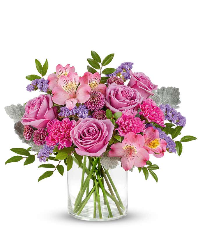 Lavender roses, pink alstroemeria, fuchsia carnations, purple button spray chrysanthemums, lavender sinuata statice, and floral greenery arranged in a short glass cylinder vase.