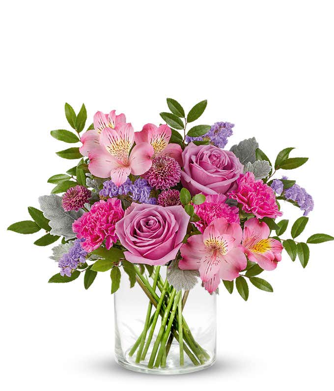 Lavender roses, pink alstroemeria, fuchsia carnations, purple button spray chrysanthemums, lavender sinuata statice, and floral greenery arranged in a short glass cylinder vase.