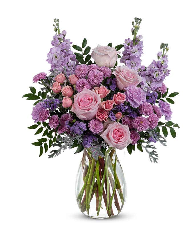 Pink roses, pink spray roses, lavender carnations, lavender stock, lavender button spray chrysanthemums, lavender sinuata statice, and floral greenery arranged in a clear glass jordan vase.