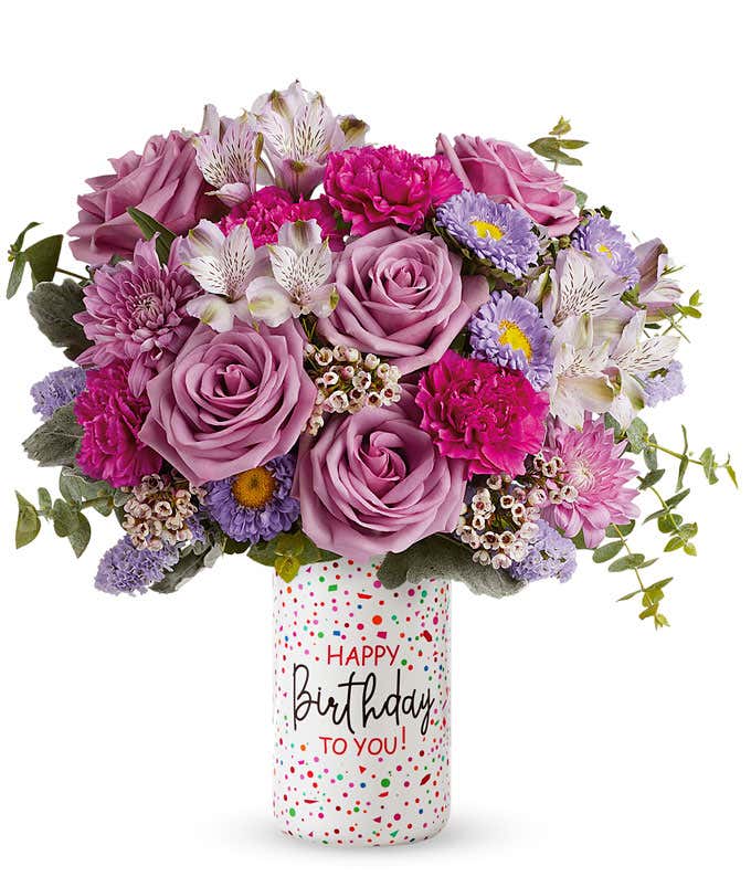 A bouquet of purple roses, asters, mums, and alstroemeria with fuchsia carnations, and greens popping out of a white cylinder vase covered in multicolor speckles and reading 