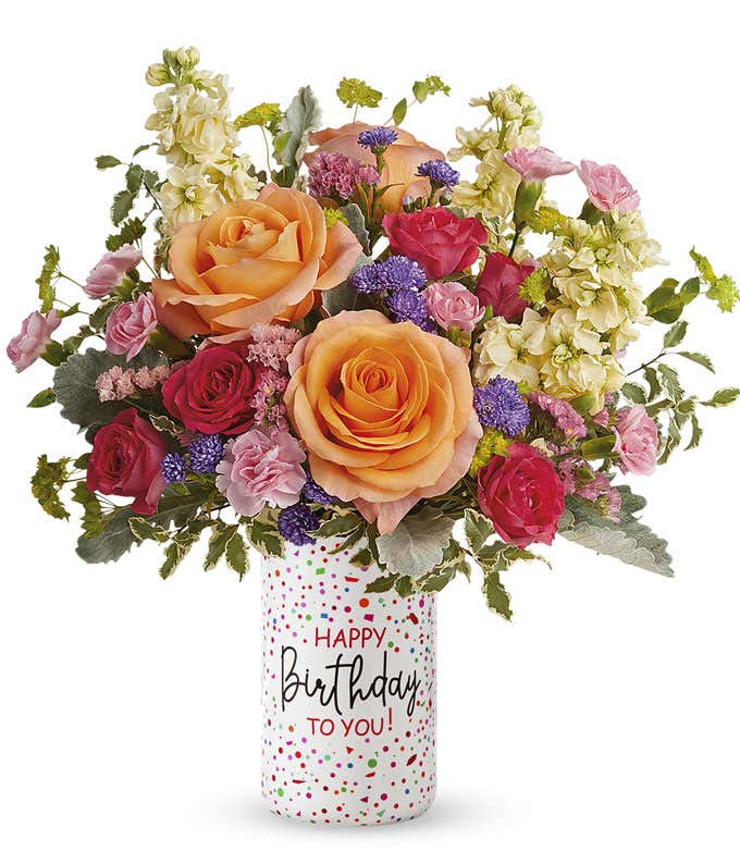 peach roses, pink mini carnations, hot pink spray roses, light yellow stock, pink sinuata statice, and purple monte casino, with greens in a white cylinder vase decorated with colorful sprinkles reading 