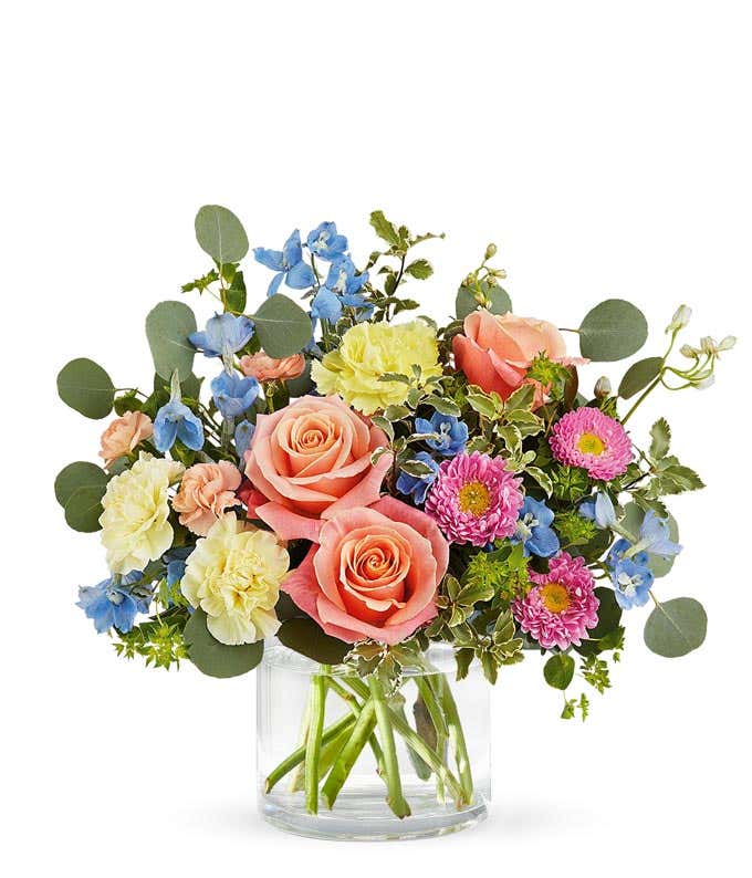 A bouquet of peach roses, yellow carnations, peach mini carnations, blue delphinium, and pink matsumoto asters with greenery in a short clear cylinder vase