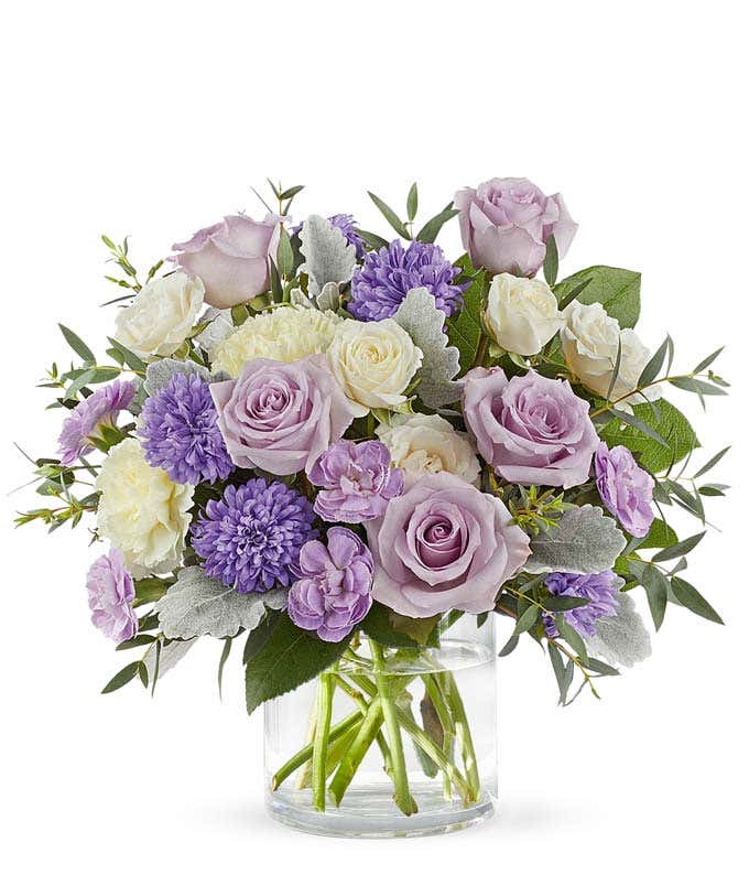 A bouquet of lavedner roses, creme spray roses, light yellow carnations, lavender mini carnations, lavender matsumoto asters, and greens in a clear short cylinder vase.