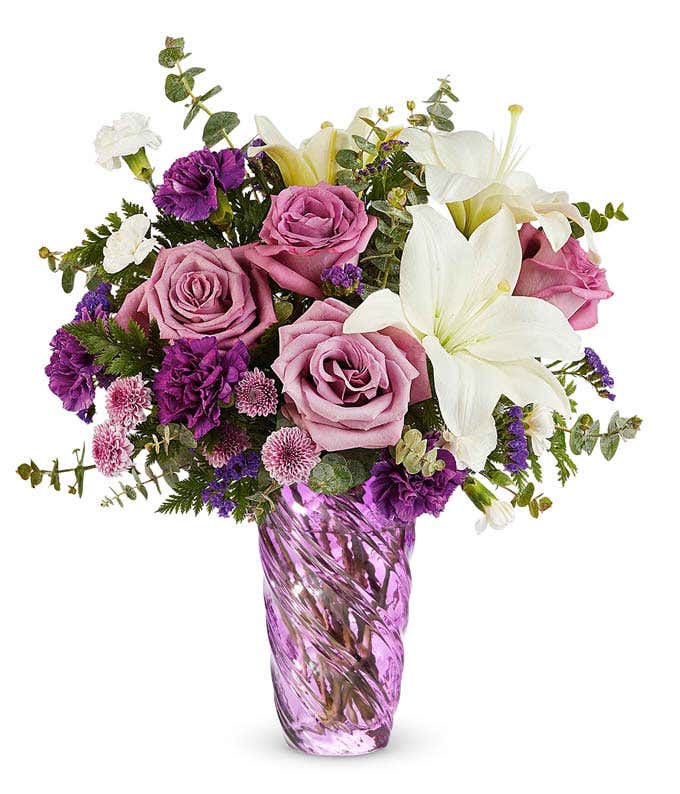 Lavender Roses, White Asiatic Lilies, Purple Carnations, Mini White Carnations, Lavender Button Poms, and Floral Greenery in a swirled lavender vase.