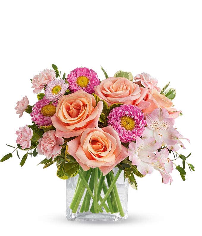 A bouquet of Peach Roses, Pink Alstroermia, Mini Pink Carnations, Pink Matsumoto Asters, in a short clear cube vase