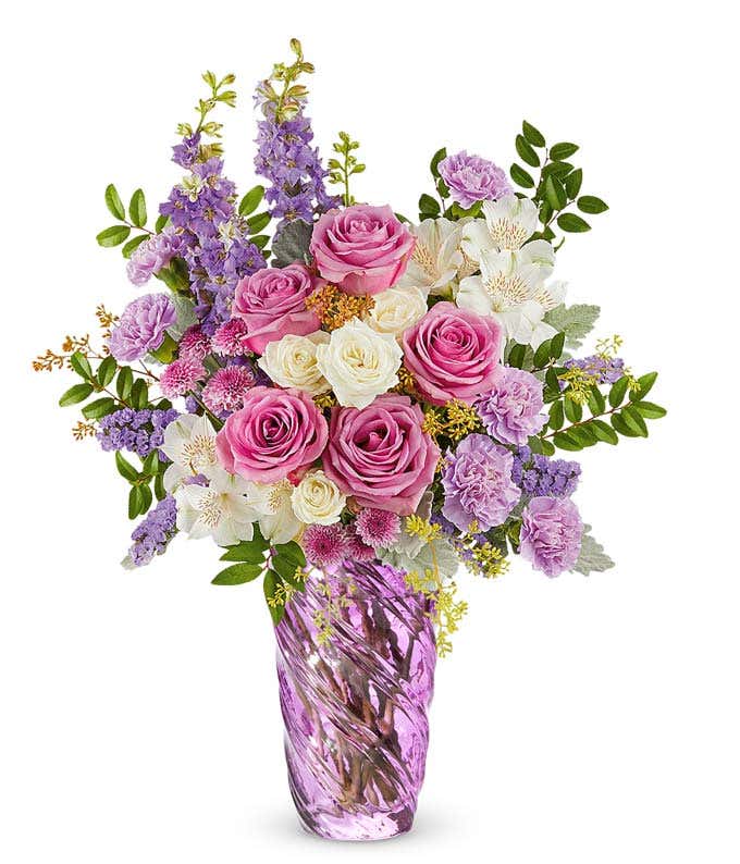 Lavender roses, white spray roses, white alstroemeria, lavender carnations, lavender larkspur, and lavender button poms with floral greenery in a swirled lavender vase