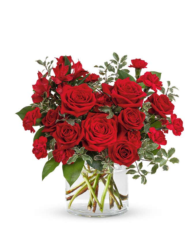 An all red bouquet with the blooms arranged in the shape of a heart consisting of roses, spray roses, alstroemeria, mini carnations, and greens in a short cylidner vase