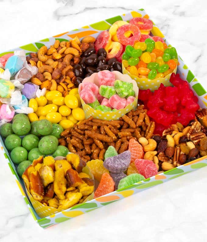 The image shows a colorful box with nine compartments, each filled with different snacks. The compartments contain assorted nuts, chocolate-covered treats, pretzels, gummy candies, fruit slices, jelly beans, licorice, hard candies, and candied corn.