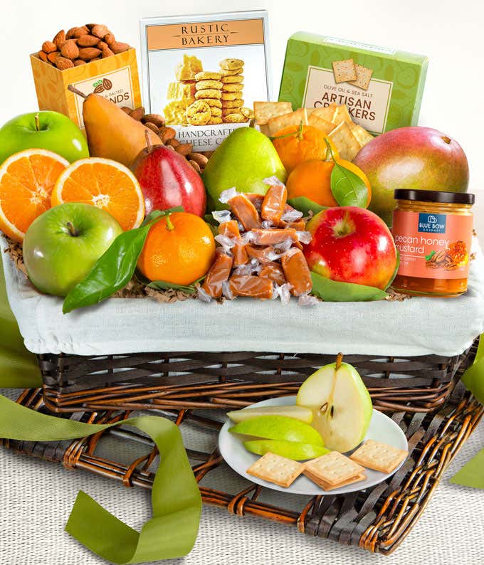 A basket filled with fresh fruits, crackers, cheese crisps, almonds, honey mustard, and assorted caramels, adorned with a green ribbon. 