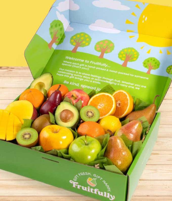 A box filled with fresh fruits including apples, oranges, pears, kiwis, avocados, and mango, nestled in green tissue paper with a colorful background.