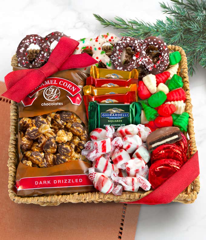 Wicker gift basket with red ribbon filled with dark chocolate drizzled caramel corn, Ghirardelli dark chocolate squares, chocolate-covered pretzels, peppermint candies, gummy Christmas trees and fish, and foil-wrapped chocolate disks.