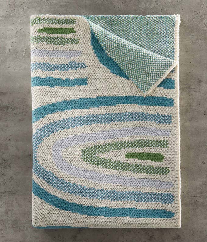  A folded blanket with a swirl pattern in green and blue shades, laid out on a grey concrete background. 