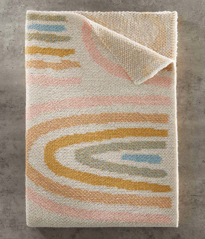 A folded blanket with a swirl pattern in pink and yellow shades, laid out on a grey concrete background.