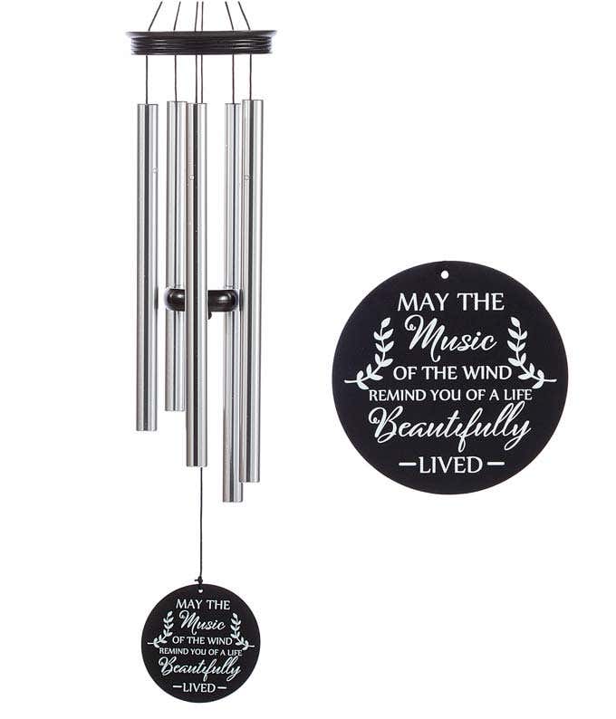 A windchime with metal rods and a wooden accents and sail. Black sail with white lettering reads may the music of the wind remind you of a life beautifully lived.
