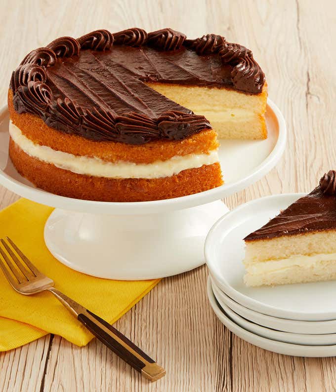 A classic Boston cream cake on a white cake stand, featuring two layers of yellow sponge cake filled with creamy vanilla custard and topped with a rich chocolate ganache. A slice is served on a stack of white plates beside a fork, ready to be enjoyed.