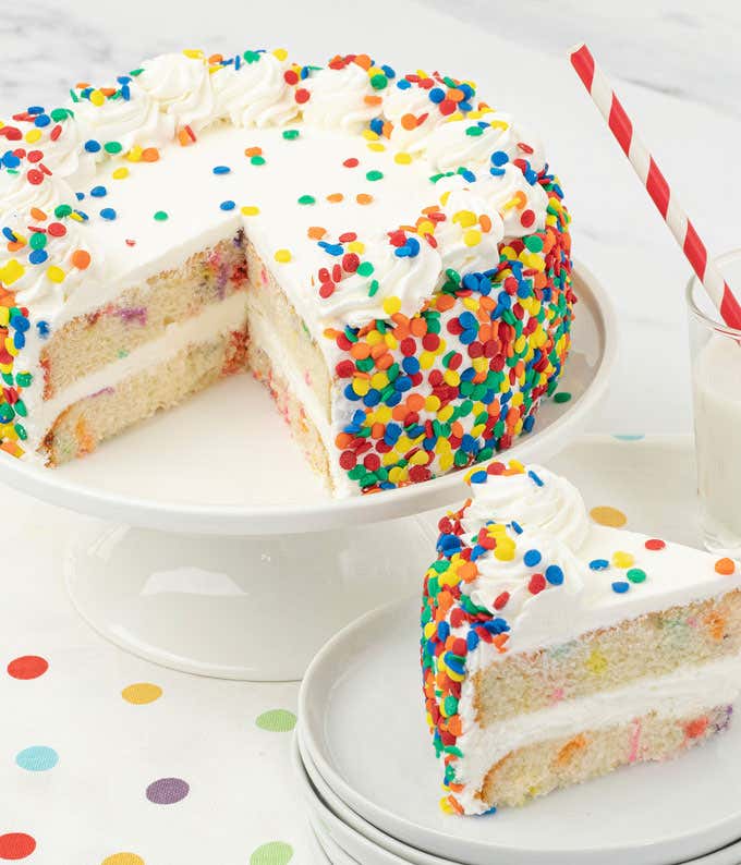  A festive confetti cake on a white cake stand, featuring two layers of vanilla sponge filled and frosted with white buttercream. The cake is decorated with colorful confetti sprinkles on the sides and top. A slice is served on stacked plates beside a gla