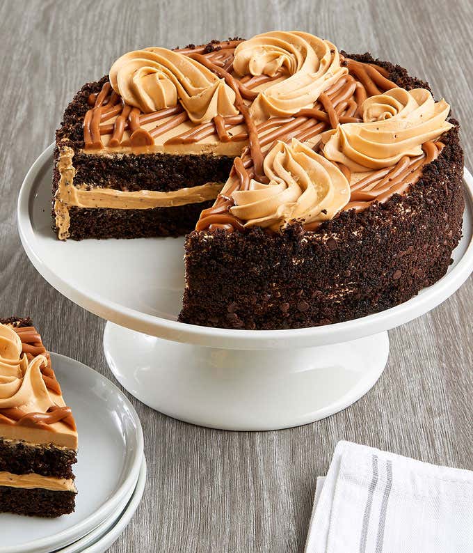 A decadent salted caramel chocolate cake on a white cake stand, featuring layers of rich chocolate cake filled and topped with smooth salted caramel buttercream. The cake is adorned with caramel drizzle and buttercream rosettes. A slice is served on stack