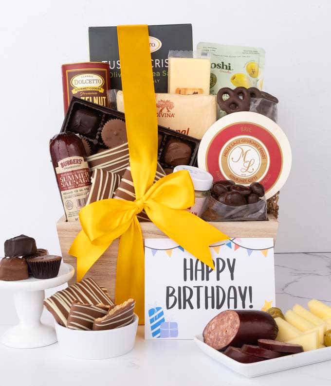 A wooden basket filled with chocolates, summer sausage, cheeses, crackers, crisps, mustard, and snacks, with displays of chocolate-covered treats, sliced sausage, and cheese styled for a Happy Birthday message card.
