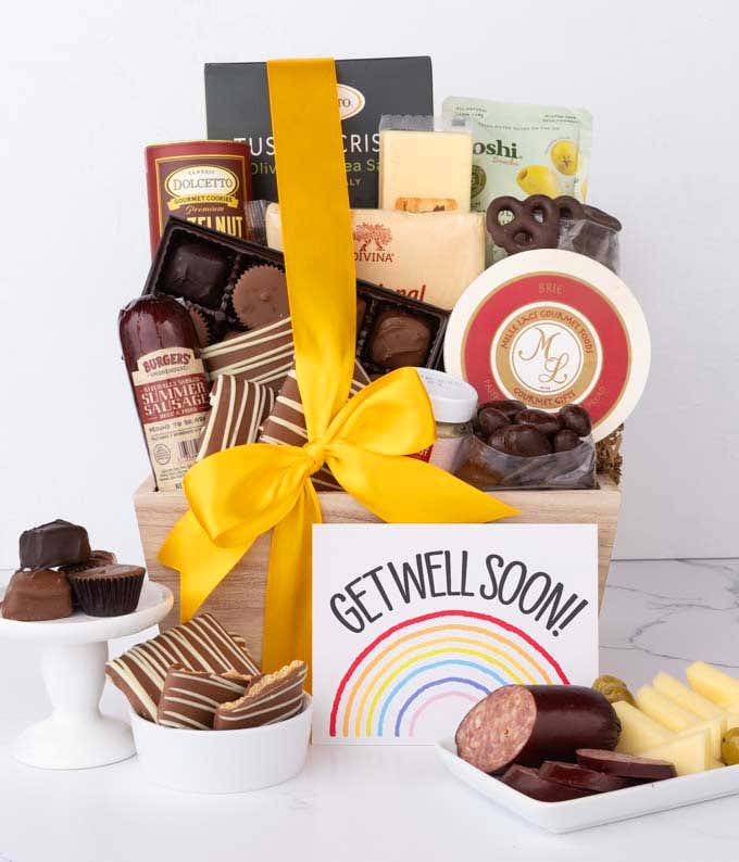 A wooden basket filled with chocolates, cheeses, sausage, crackers, crisps, and snacks, displayed with a rainbow Get Well Soon card, chocolate treats, and sliced cheese and sausage.