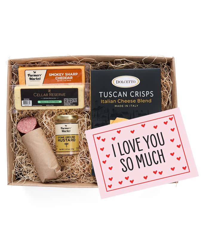 A gift box filled with two different hard cheeses, a small jar of stone-ground mustard, gourmet crackers, a summer sausage, and a pink card with hearts that reads 'I Love You So Much'