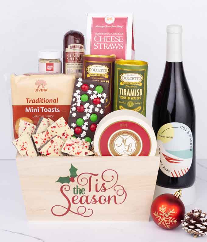 A wooden basket with the words Tis the Season, filled with sausage, cheese, mustard, cookies, wafers, and holiday bark, paired with a bottle of Hill & Harbor Pinot Noir beside a red ornament and pinecone.