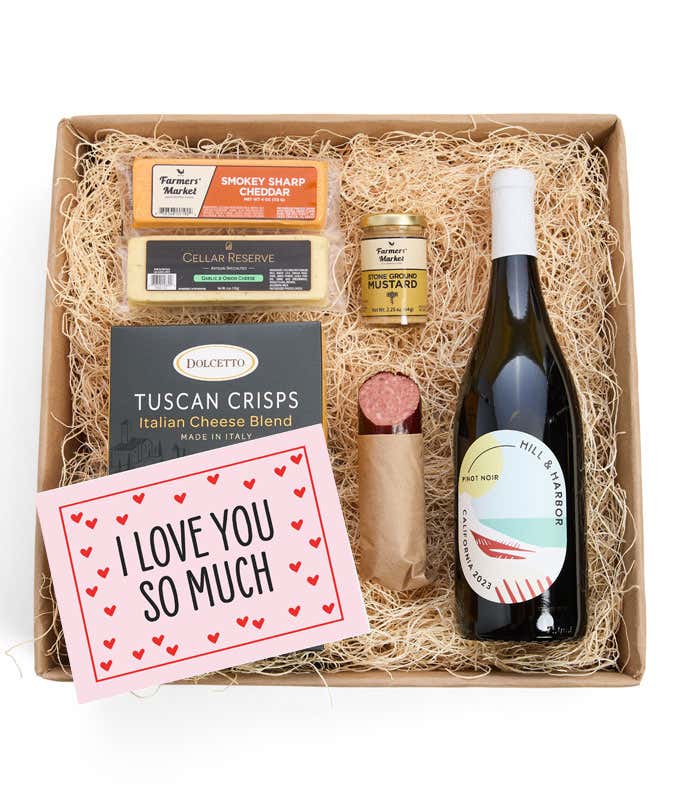 A beautifully arranged gift box featuring a bottle of Hill & Harbor Pinot Noir, two hard cheeses, a jar of stone-ground mustard, gourmet crackers, savory summer sausage, and a pink card with hearts that reads 'I Love You So Much'