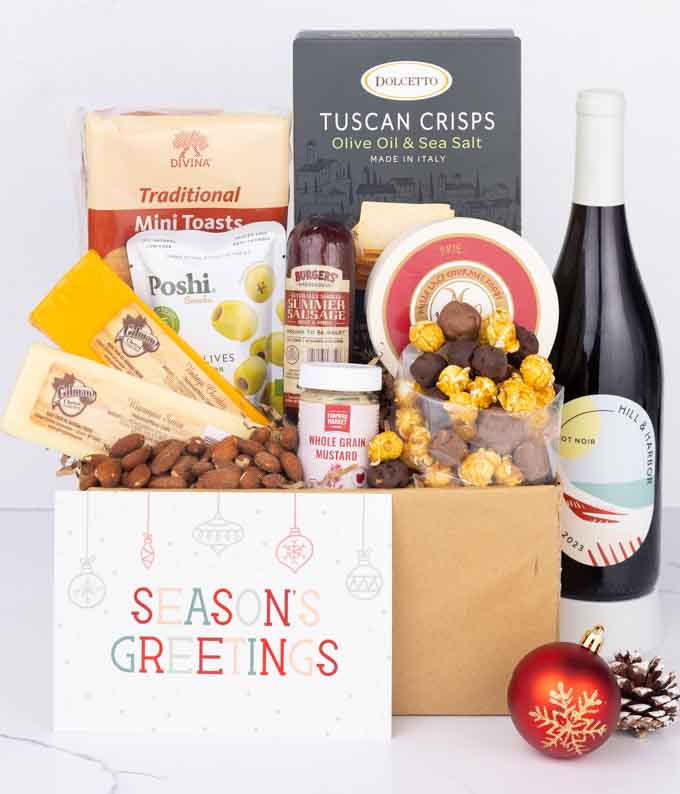 A charcuterie gift set with 3 cheeses, Tuscan crisps, focaccia crackers, olives, mustard, smoked almonds, chocolate-covered popcorn, summer sausage, a bottle of Pinot Noir, and a Season’s Greetings card.
