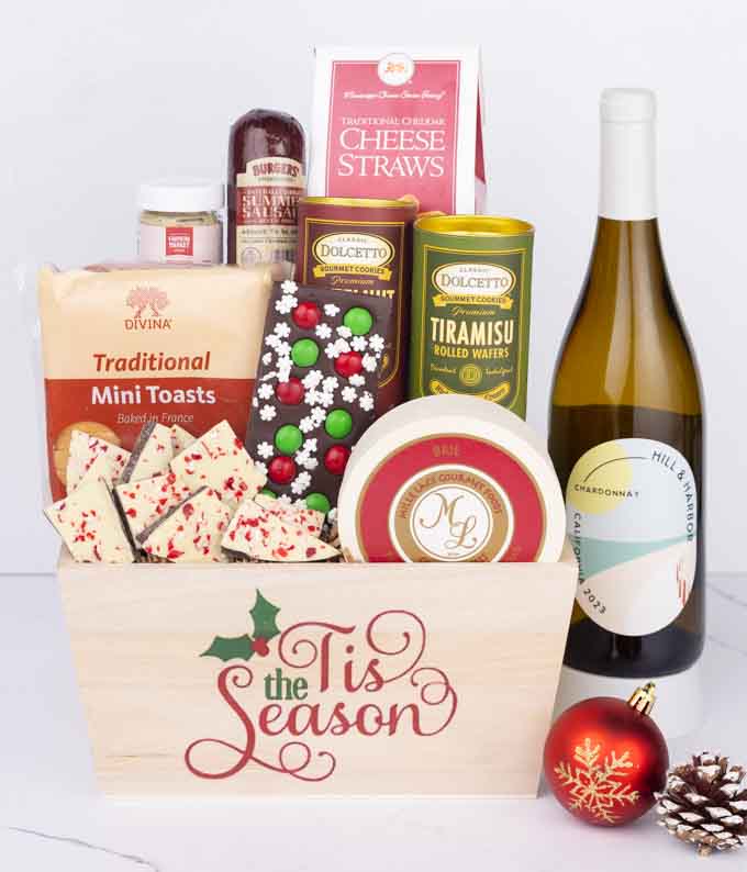 A wooden basket with the words Tis the Season, filled with sausage, cheese, mustard, cookies, wafers, and festive bark, paired with a bottle of Hill & Harbor Chardonnay, next to a red ornament and pinecone.