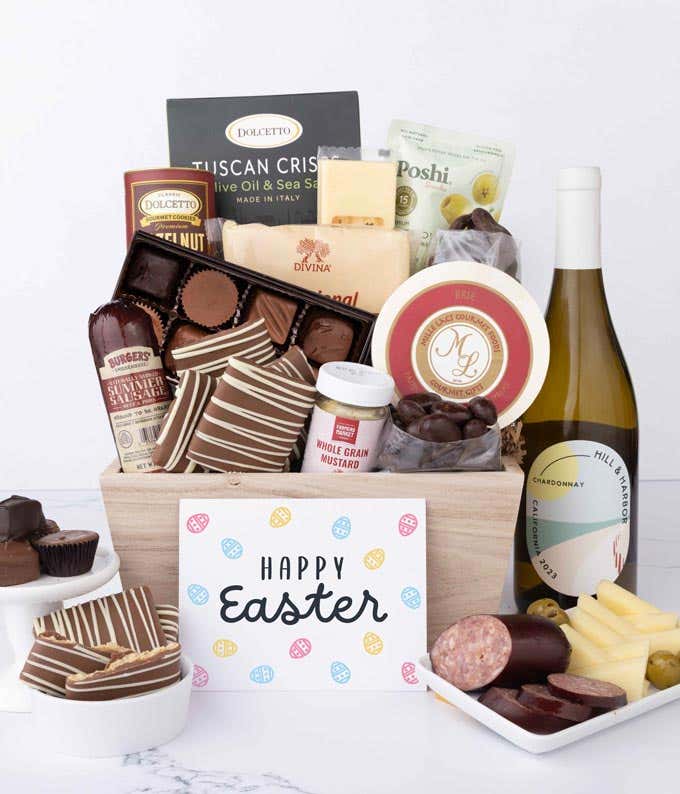 A wooden crate filled with gourmet snacks including sausage, assorted cheeses, crackers, olives, mustard, chocolate almonds, pretzels, graham crackers, assorted chocolates, and wafers, with a bottle of wine and an Easter card.