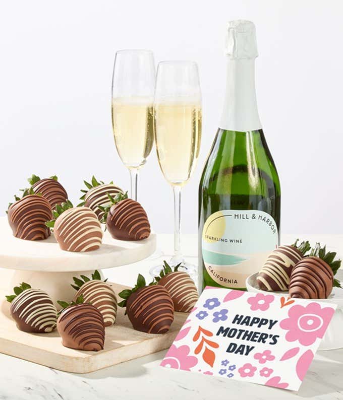 Hill & Harbor sparkling wine bottle displayed with two glasses and assorted chocolate-covered strawberries on trays, styled with a floral Happy Mother’s Day card for a festive wine and dessert gift.