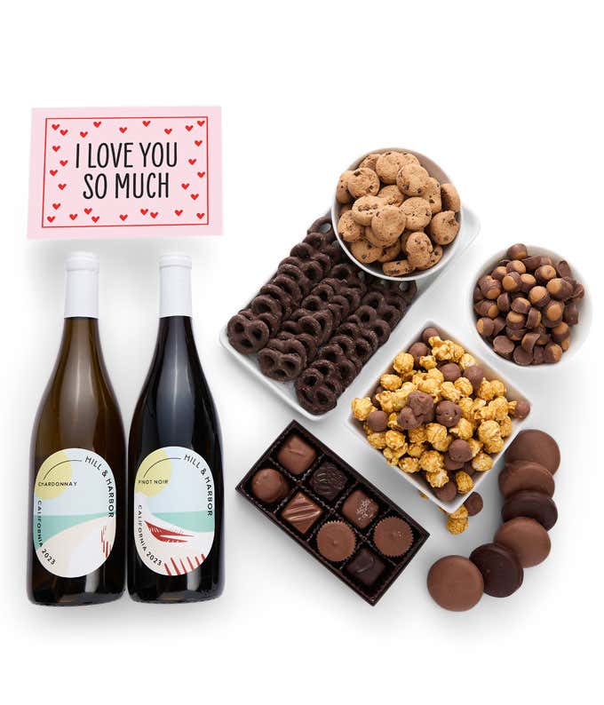 A gift box overflowing with treats including an assortment of chocolate-covered items like oreoes, almonds graham crackers and pretzels along with red velvet cookies, hazelnut wafers, an 8 piece box of chocolates, 2 bottles of wine and an I Love You card.