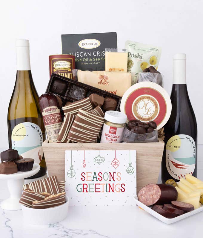 A wooden basket filled with chocolates, summer sausage, cheeses, crackers, crisps, mustard, and snacks, with displays of chocolate treats, sliced sausage, olives, and cheese arranged with 2 bottles of wine and Season's Greetings message.