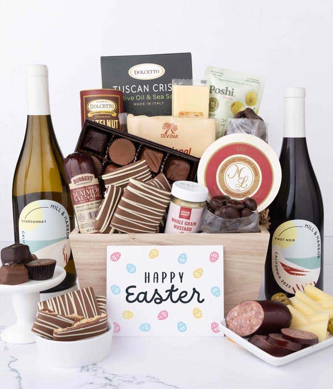 A wooden crate filled with gourmet snacks including sausage, assorted cheeses, crackers, olives, mustard, chocolate almonds, pretzels, graham crackers, assorted chocolates, and wafers, with 2 bottles of wine and an Easter card.