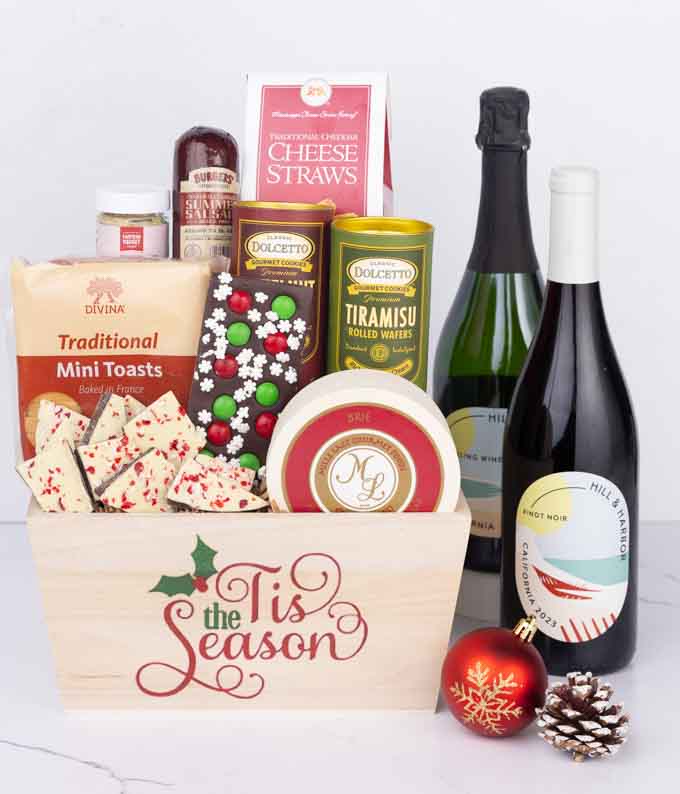 A wooden basket with the words Tis the Season, filled with sausage, cheese, mustard, cookies, wafers, and holiday bark, paired with bottles of Hill & Harbor Pinot Noir and Sparkling Wine, accented by a red ornament and pinecone.