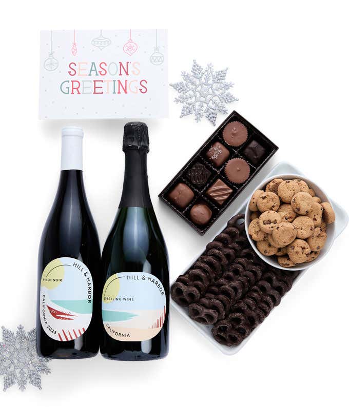 A holiday gift set with two bottles of Hill & Harbor wine (Pinot Noir and Sparkling Wine), a box of assorted chocolates, a tray of chocolate-covered pretzels, a bowl of mini chocolate chip cookies, and a Season's Greetings message card.