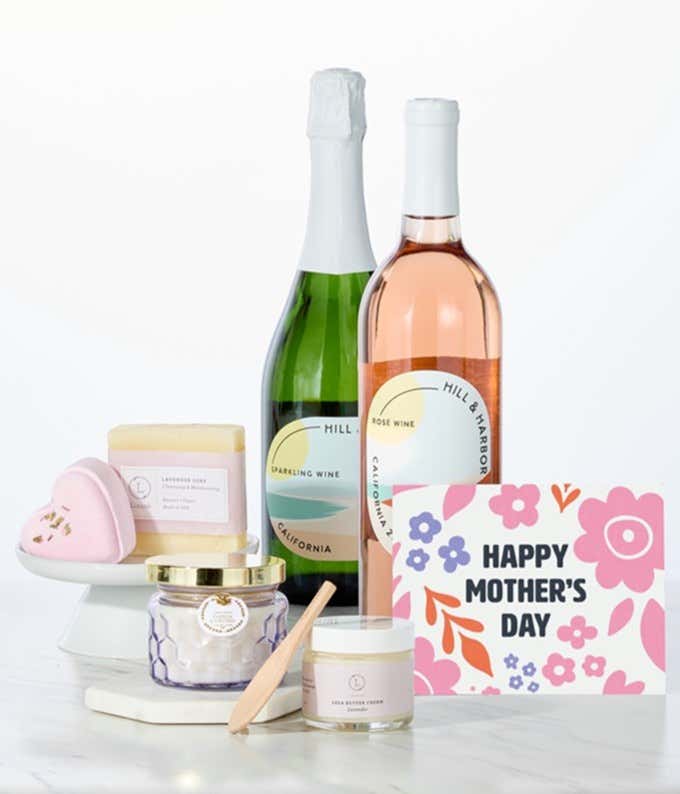 Two bottles of Hill & Harbor California wine paired with spa essentials including artisan lavender soap, a heart-shaped shower steamer, shea butter cream with wooden scoop, and a candle arranged in a clean display with a Mother's Day card.