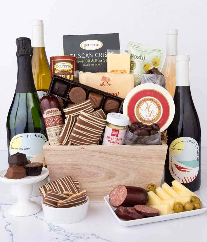 Wooden crate filled with gourmet food and 4 bottles of wine, including Chardonnay, Pinot Noir, Sparkling Wine, and Rosé, alongside artisan summer sausage, creamy cheeses, assorted chocolates, striped chocolate grahams, cookies, crisps, mustard, and olives