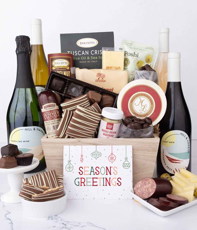 A wooden basket filled with chocolates, summer sausage, cheeses, crackers, crisps, mustard, and snacks, with displays of chocolate treats, sliced sausage, olives, and cheese arranged with 4 bottles of wine and Season's Greetings message.