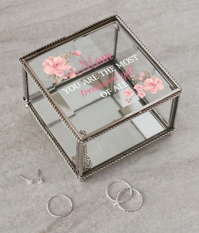 Glass jewelry box, with a plated silver braided metal trim, and a quote that reads: Mom you are the most treasured gift of all, surrounded by pink flowers
