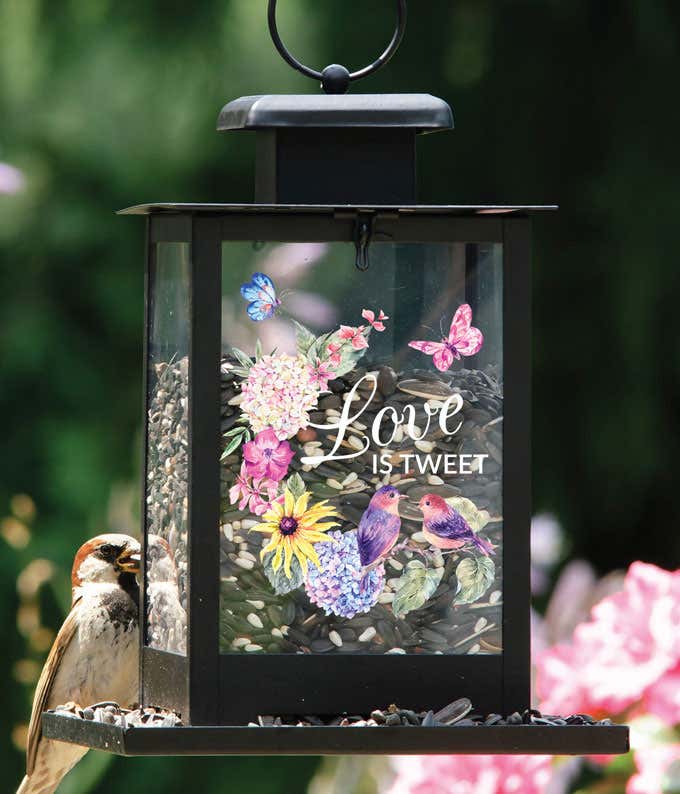 Black lantern-style bird feeder with a floral design, featuring colorful flowers, birds, butterflies, and the phrase Love is Tweet