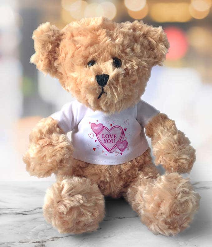 Light brown teddy bear with a fluffy texture, wearing a white shirt featuring pink heart designs and the text 