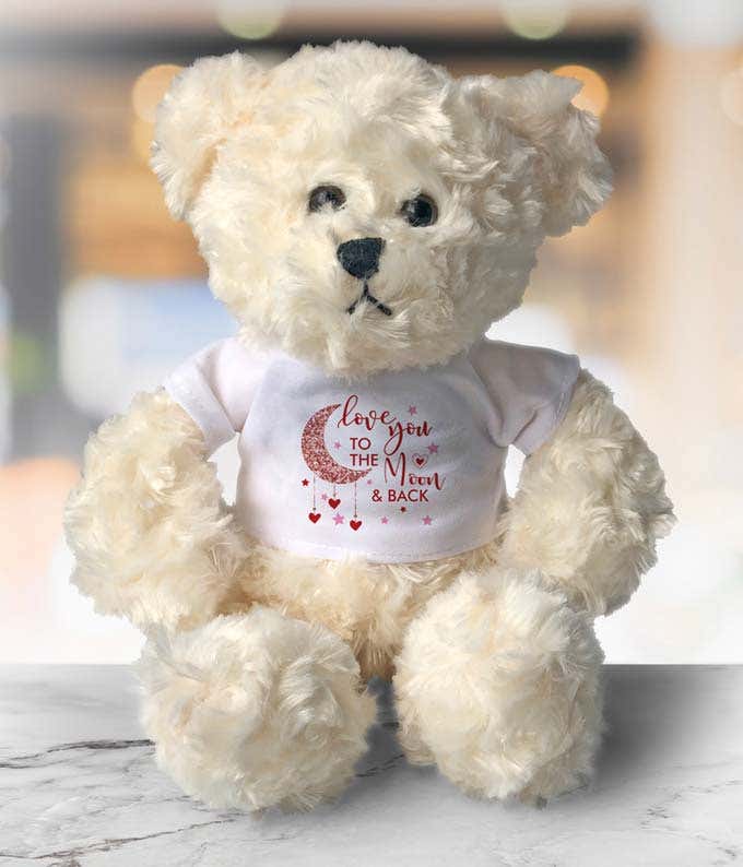 Cream-colored teddy bear with soft, fluffy fur, wearing a white shirt featuring a crescent moon design, pink hearts, and the message Love You to the Moon & Back.