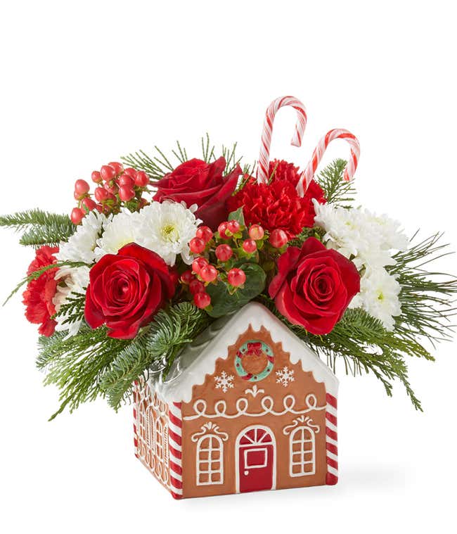 Festive holiday arrangement with red roses, red carnations, white poms, red hypericum berries, and seasonal greenery, accented with candy canes. Displayed in a gingerbread house container with white icing details and candy decorations.