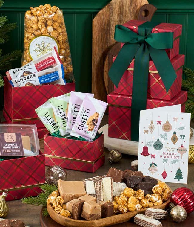 A festive pink and green argyle gift tower with a white bow, filled with chocolate peanut butter fudge, Cubetti wafer cookies, dark chocolate sea salt caramels, and caramel popcorn. A delightful holiday treat assortment perfect for gifting.