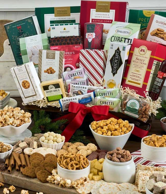  Large gift basket filled with an assortment of gourmet snacks including cheeses, crackers, sausages, popcorn, chocolates, nuts, cookies, and various specialty treats, arranged on wooden boards and dishes with festive decorations.