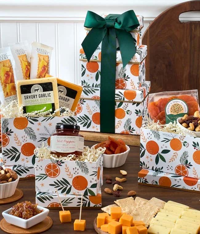 A stylish gift tower with a green ribbon, featuring three decorative boxes filled with savory garlic and sharp cheddar cheeses, sourdough flatbreads, dried apricots, mixed nuts, hot pepper jelly, and cheese cubes with crackers.