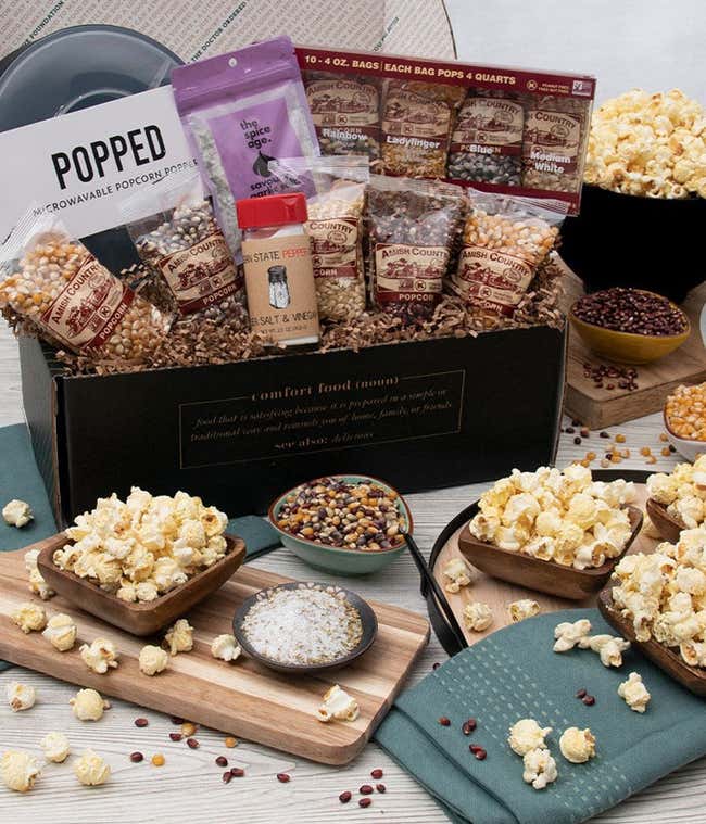 Gift box with 10 bags of popcorn kernels, a microwave popcorn popper, salt &amp; vinegar seasoning, bag of garlic seasoning. In front of the gift basket are 4 small bowls of popped popcorn, 3 small bowls of kernels on a grey wooden table.