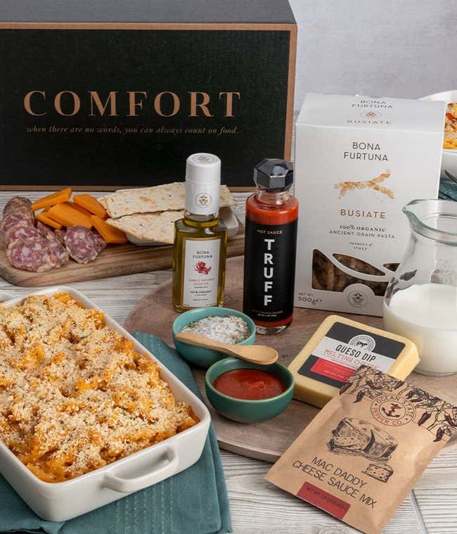 Gift box on a table with salami, cheddar cheese, and crackers on one tray, another tray with a box of pasta, bowls of seasoning, bottles of olive oil &amp; hot sauce, a block of cheese, and a packet of mac daddy sauce. On the table is also a baking dish with 