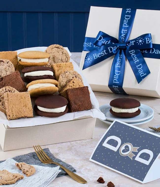 The image features a box labeled 'Best Dad Ever' filled with an assortment of cookies and brownies. The treats are arranged on a marble and wood cutting board, with additional cookies on a small plate nearby.